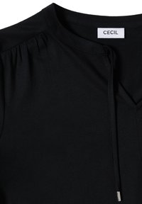 T-shirt noir en tissu doux, col rond, plis subtils aux épaules et cordon de serrage au col avec embout métallique. Étiquette de la marque : CECIL.