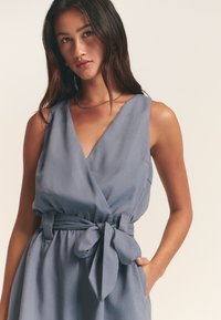Světle modré zavinovací šaty vyrobené z jemného materiálu, s výstřihem do V, širokými ramínky, opaskem na zavázání v pase a bočními kapsami.