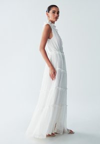 Abito maxi bianco con scollatura arricciata e design a strati; realizzato in tessuto leggero con una sottile texture e elementi trasparenti.