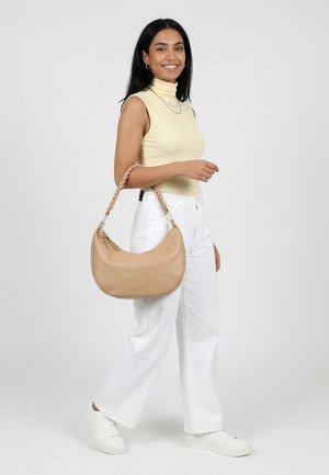 Vrouw draagt een mouwloze beige coltrui en witte broek, houdt een beige handtas met gedraaide handgreep aan haar arm vast, lacht naar de camera.