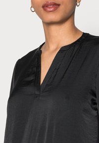 Svart blus med V-ringning, strukturerade axlar och en slät, blank yta. Detaljer inkluderar en diskret knapplist.