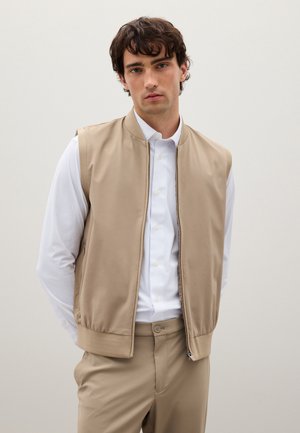 Joven con cabello rizado oscuro que lleva un chaleco beige sin mangas con cremallera, una camisa blanca de manga larga y pantalones beige, de pie frente a un fondo sencillo.