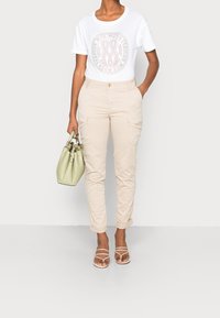 Vit T-shirt med en cirkulär logotyp, beige cargo-byxor och en ljusgrön handväska. Sandaler med remmar fullföljer looken. Enkel, funktionell design.