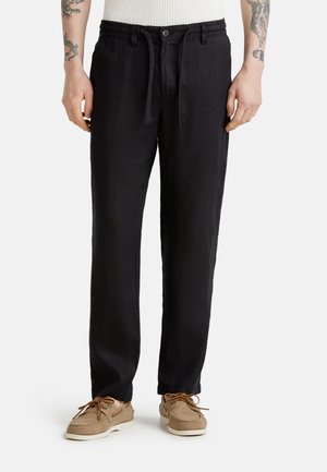 Person trägt schwarze Kordelhose, beige Mokassinschuhe und ein weißes strukturiertes, ärmelloses Oberteil, steht vor weißem Hintergrund.