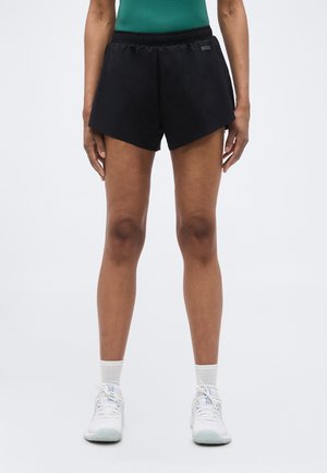 Osoba ubrana w czarne sportowe szorty, białe skarpetki do kostek, białe trampki i zielony top, stojąca na tle jednolitego jasnego tła.