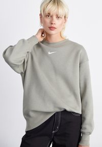 Sweatshirt cinza com gola redonda e mangas longas, apresentando um pequeno logotipo branco da Nike no peito. Usado com calças pretas, exibindo um corte descontraído.