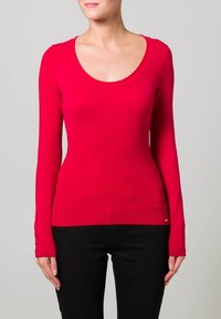 Rode, geribbelde top met lange mouwen en een ronde halslijn, met een getailleerde pasvorm. Bevat subtiele accent details bij de manchetten. Gecombineerd met zwarte broek.