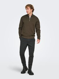 Pull vert olive foncé avec une fermeture éclair sur le devant jusqu'au quart, fabriqué en tissu tricoté texturé, associé à un pantalon gris foncé ajusté et des bottes noires.