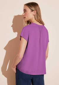 Top a maniche corte in tessuto viola brillante, con scollatura rotonda e dettaglio plissettato sul retro, abbinato a pantaloni blu scuro.