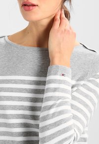 Maglione a righe grigie e bianche con scollo a barca, texture a maglia e accentuazione del marchio visibile sulla manica.