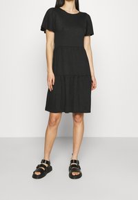 Robe noire à manches courtes avec une jupe à volants, confectionnée dans un tissu lisse. Elle présente un col rond et arrive juste au-dessus des genoux.