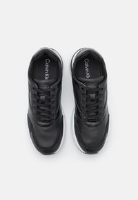 Baskets en cuir noir à finition lisse, avec lacets, semelle blanche et un logo discret sur le côté. Des accents texturés rehaussent le design.