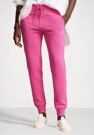 Persona con pantalones deportivos de color rosa brillante con cintura de cordón, zapatillas blancas y una camisa blanca parcialmente metida, de pie frente a un fondo liso.