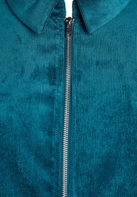 Zielona dżinsowa kurtka na zamek błyskawiczny z wyraźną żeberkową teksturą i srebrnym zamkiem. Klatka z kołnierzem podkreśla zorganizowany wygląd.