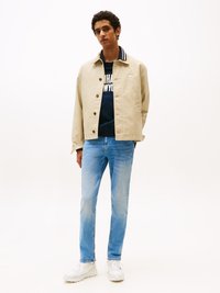 Beige Jacke mit Knopfdetails und gestreiftem Kragen, dunkelblauem Sweatshirt mit Aufschrift, hellblauen Jeans und weißen Sportschuhen.