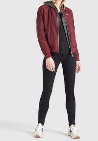 Vínová bomber bunda s elastickými manžetami, šedou kapucí a předním zipem; doplněná o černé legíny a bílé tenisky s modrými prvky.