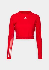 Czerwony, długi crop top z elastycznego materiału. Posiada stójkę, siateczkowe akcenty na rękawach oraz biały logo z trzema paskami na lewym rękawie.