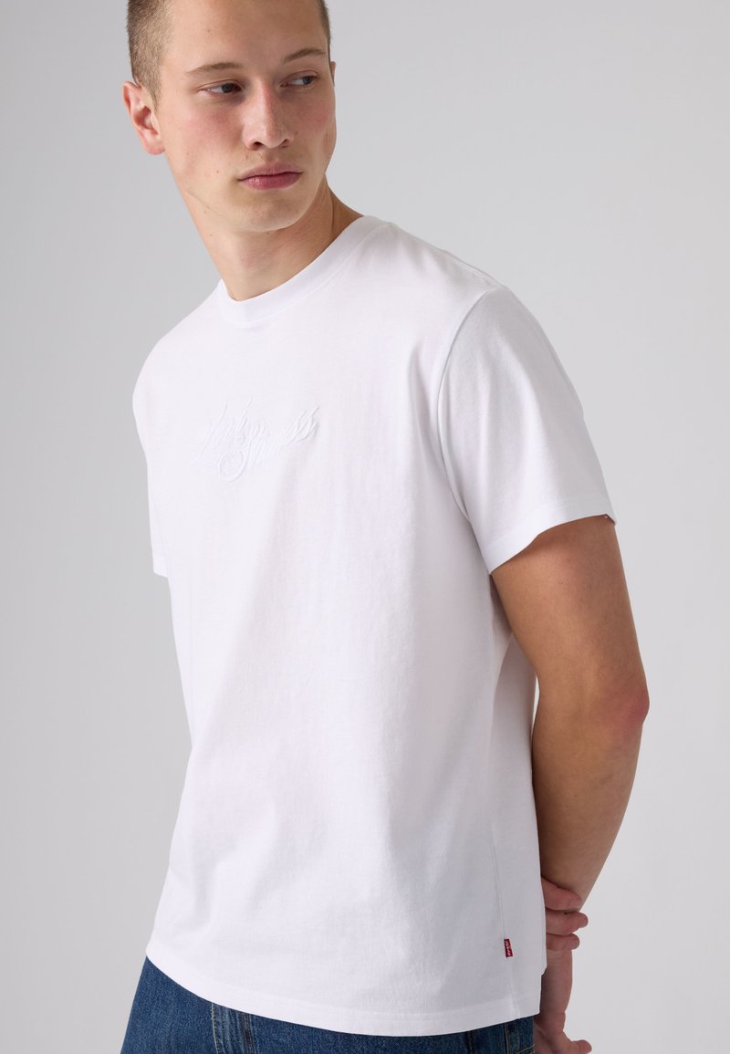 Young man wearing a plain white short-sleeve t-shirt and blue jeans, looking to his left with hands behind his back.
