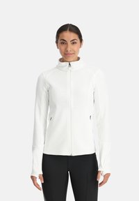 Veste blanche zippée en tissu à texture côtelée, avec col montant, manches longues et poches latérales, associée à un pantalon noir.