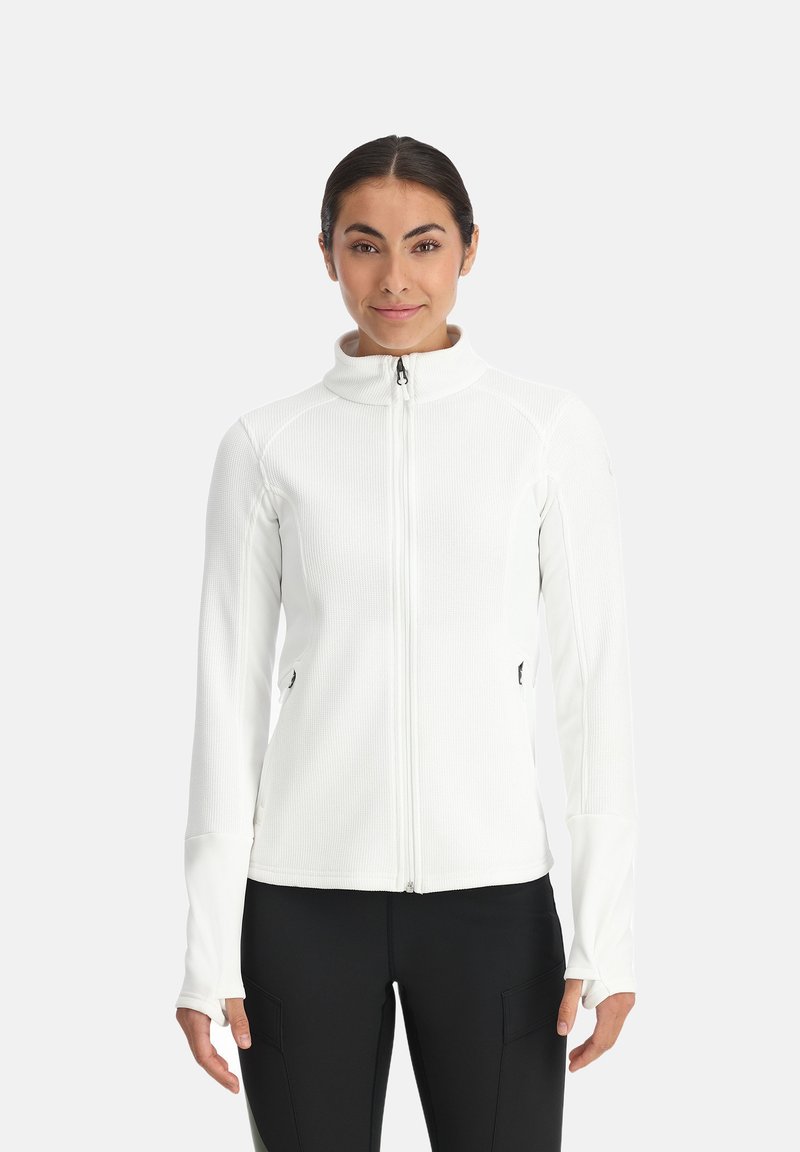Veste blanche zippée en tissu à texture côtelée, avec col montant, manches longues et poches latérales, associée à un pantalon noir.