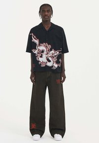 Camisa negra de manga corta con un gráfico de dragón en blanco y rojo; combinada con pantalones holgados de color verde oscuro con acentos rojos y calzado blanco.