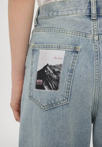 Jeans de denim azul claro com um patch gráfico de montanha em preto e branco no bolso traseiro, com costuras brancas e o texto "Moschino Jeans".