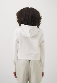 Hanorac alb scurt, cu glugă ajustabilă, având un logo subtil pe spate. Material moale, manșete cu tiv, asortat cu pantaloni scurți de culoare deschisă.