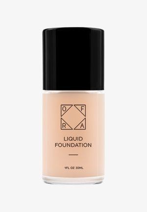 Fond de teint dans un flacon en verre transparent, liquide beige, avec un capuchon noir élégant. L'étiquette présente le nom de la marque et les détails du produit en impression noire.