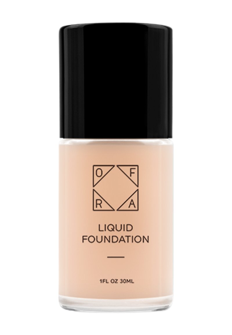 Vloeibare foundation in een transparante glazen fles, beige vloeistof, met een elegante zwarte dop. Het etiket bevat de merknaam en productdetails in zwarte druk.