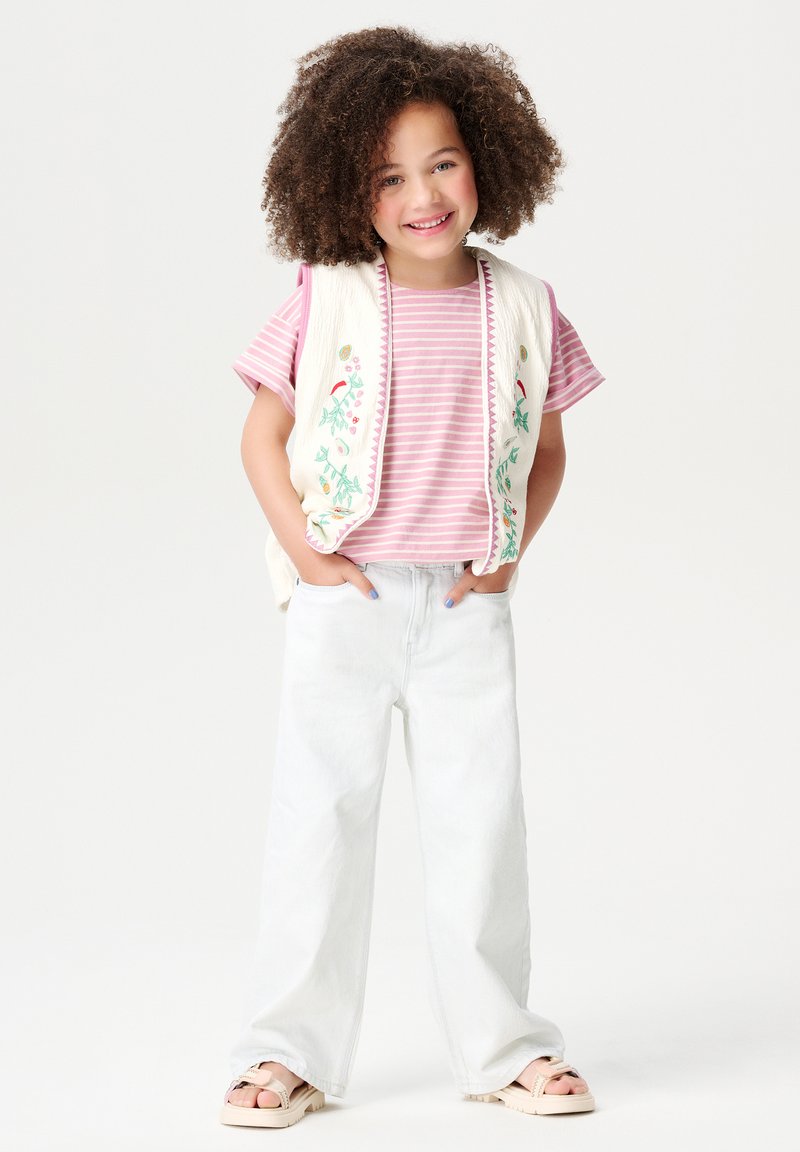 Witte geborduurde vest over een roze en witte gestreepte shirt, gecombineerd met lichtdenim wijde broek en beige sandalen. Krullend haar dat het gezicht omlijst.