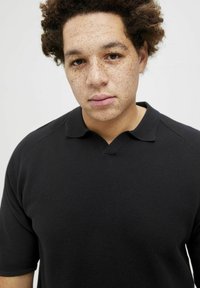 Juodas polo marškinėlis iš tekstūruoto audinio, turintis klasikinį apykaklę ir trumpas rankoves, demonstruojamas ant modelio su šviesiomis dėmėmis.