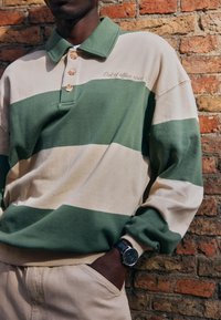 Homme portant un polo à manches longues rayé vert et beige et un pantalon beige, debout contre un mur en briques avec une main dans la poche.