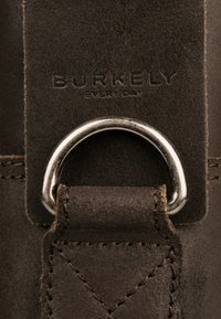 Étiquette en cuir brun avec le texte embossé "BURKELY EVERY DAY", dotée d'un anneau en métal argenté et d'un détail de couture visible.