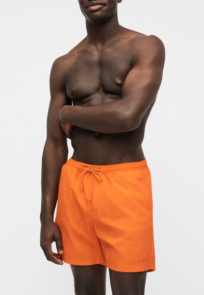 Homme torse nu à la peau foncée portant un short de bain orange vif noué à la taille, debout devant un fond clair uni.