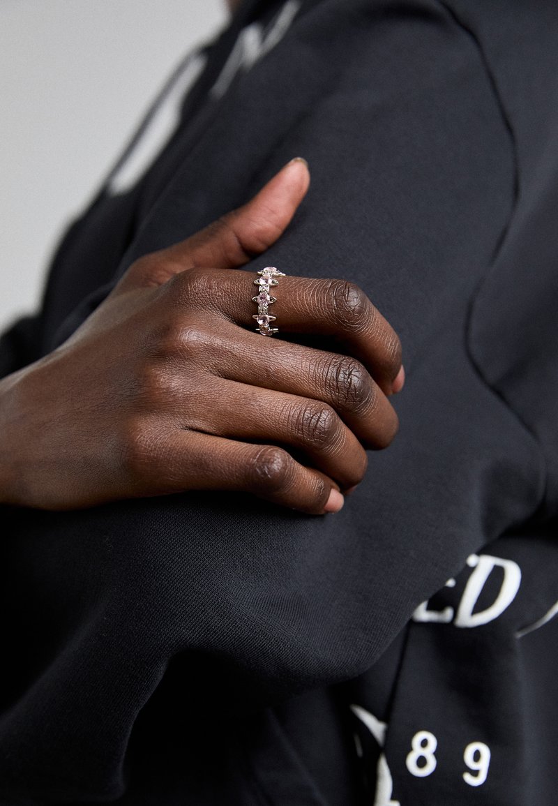 Hopeasormus, jossa on vuorotellen kirkkaat ja vaaleanpunaiset kivet, ja teksturoitu rengasosa. Käsi lepää mustaa kangasta vasten.