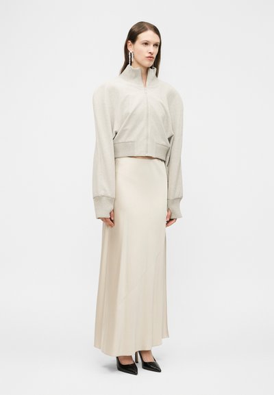 Femme portant un pull beige court avec fermeture éclair et manches bouffantes, une longue jupe crème, des talons pointus noirs et des boucles d'oreilles pendantes, debout.