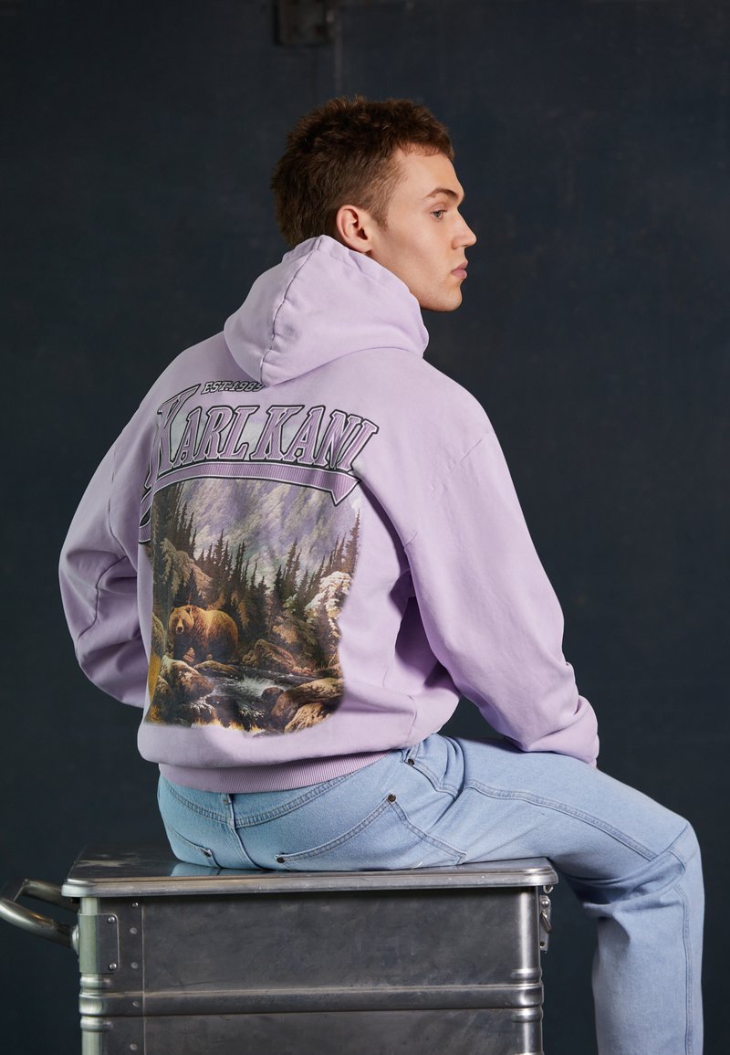 Felpa viola con un motivo naturale che raffigura un orso e degli alberi sul retro. Abbinata a jeans azzurri chiari, la modella è seduta su una superficie metallica.