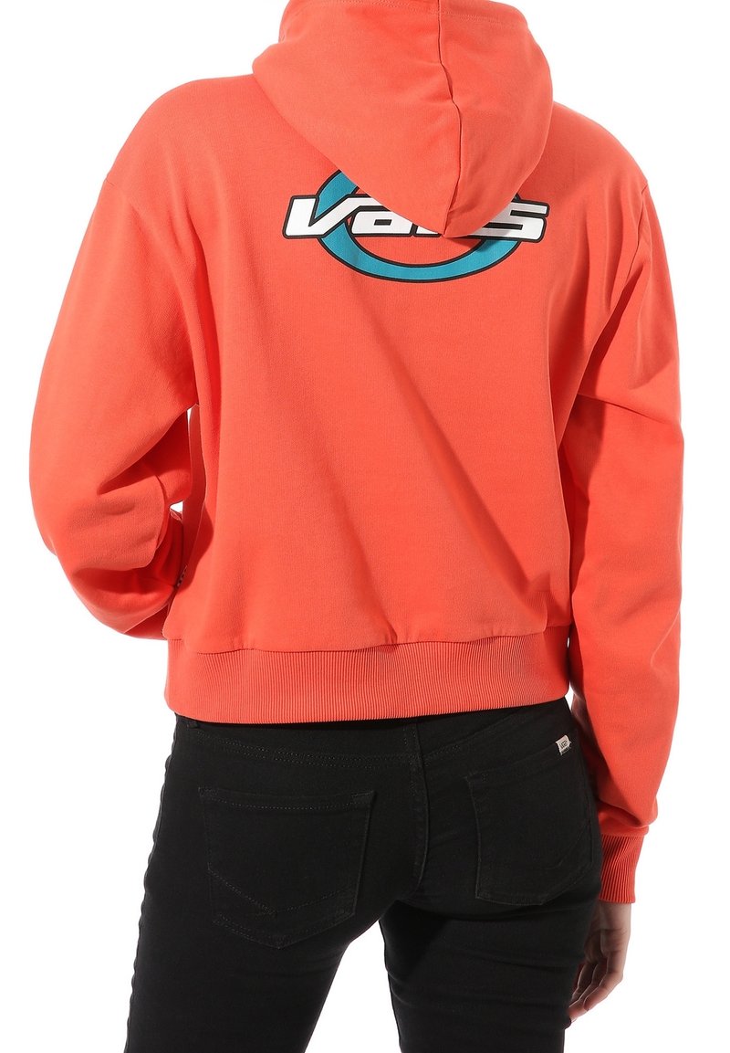 Persona che indossa un vivace hoodie arancione con il logo Vans blu e bianco sulla parte superiore della schiena, abbinato a jeans neri.
