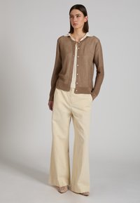Mujer con un cárdigan tejido marrón abotonado sobre una camisa blanca con cuello, pantalones anchos crema y zapatos beige tejidos, de pie contra un fondo claro.
