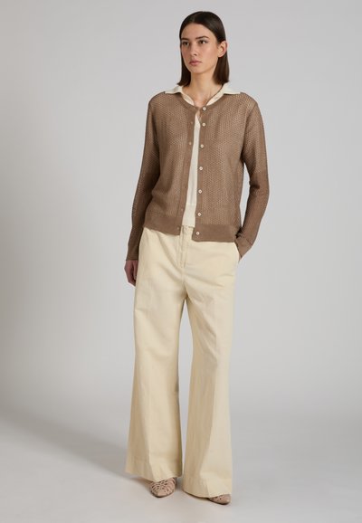 Mujer con un cárdigan tejido marrón abotonado sobre una camisa blanca con cuello, pantalones anchos crema y zapatos beige tejidos, de pie contra un fondo claro.