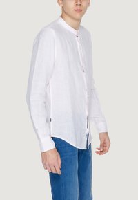 Camicia bianca in lino con colletto alla mandarino, pattina dei bottoni e maniche lunghe, caratterizzata da un orlo curvo e un design minimalista.