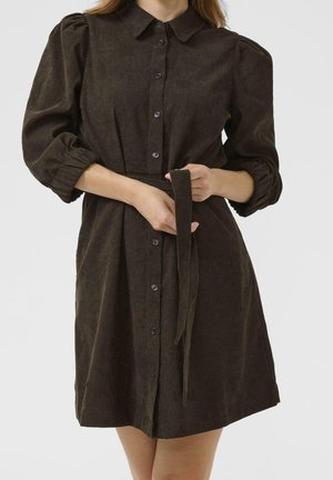 Femme portant une robe boutonnée marron foncé à manches bouffantes, tenant la ceinture nouée devant un fond clair uni.