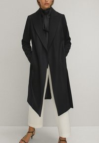 Manteau long noir avec un col cranté et un design à l'avant ouvert, fabriqué en tissu lisse. Associé avec une écharpe sombre et un pantalon large crème.