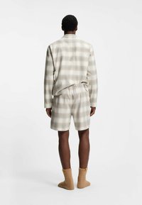 Mann von hinten zu sehen, der ein beiges und graues kariertes Langarmhemd mit passenden Shorts und flauschigen braunen Socken vor einem weißen Hintergrund trägt.