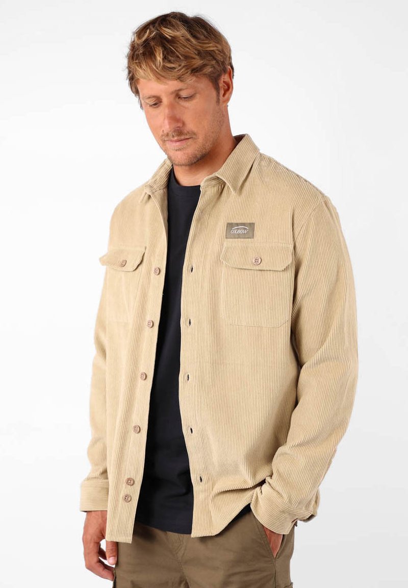 Chemise en velours côtelé beige avec bouton devant, deux poches poitrine et une patch de marque. Portée par-dessus une chemise noire, avec une coupe décontractée et une texture côtelée.