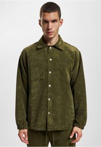 Olive green corduroy jacket featuring a collared neck, snap buttons, and two front pockets. Textured fabric with a slightly shiny finish.