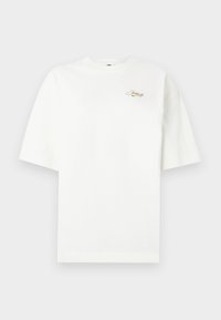 Λευκό oversized κοντομάνικο μπλουζάκι με μικρό χρυσό λογότυπο "Just Do It" στο άνω αριστερό μέρος του στήθους σε απλή φόντο.