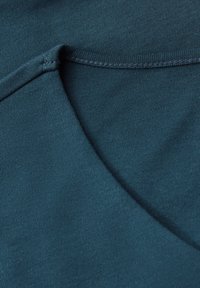 Detailní záběr na tyrkysovou látku, kde je vidět podrobné šití a struktura složeného bavlněného oděvu.