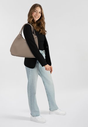Beige hobo tas met een gebogen vorm en een zachte textuur, op de schouder gedragen. Zwart langemouwtop, lichtblauwe spijkerbroek, witte sneakers.