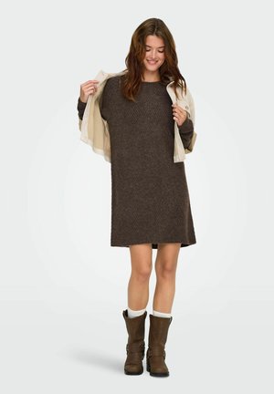 Vestido de punto marrón con un corte holgado y un patrón texturizado, combinado con botas beige y calcetines blancos, mientras sostiene una chaqueta crema.